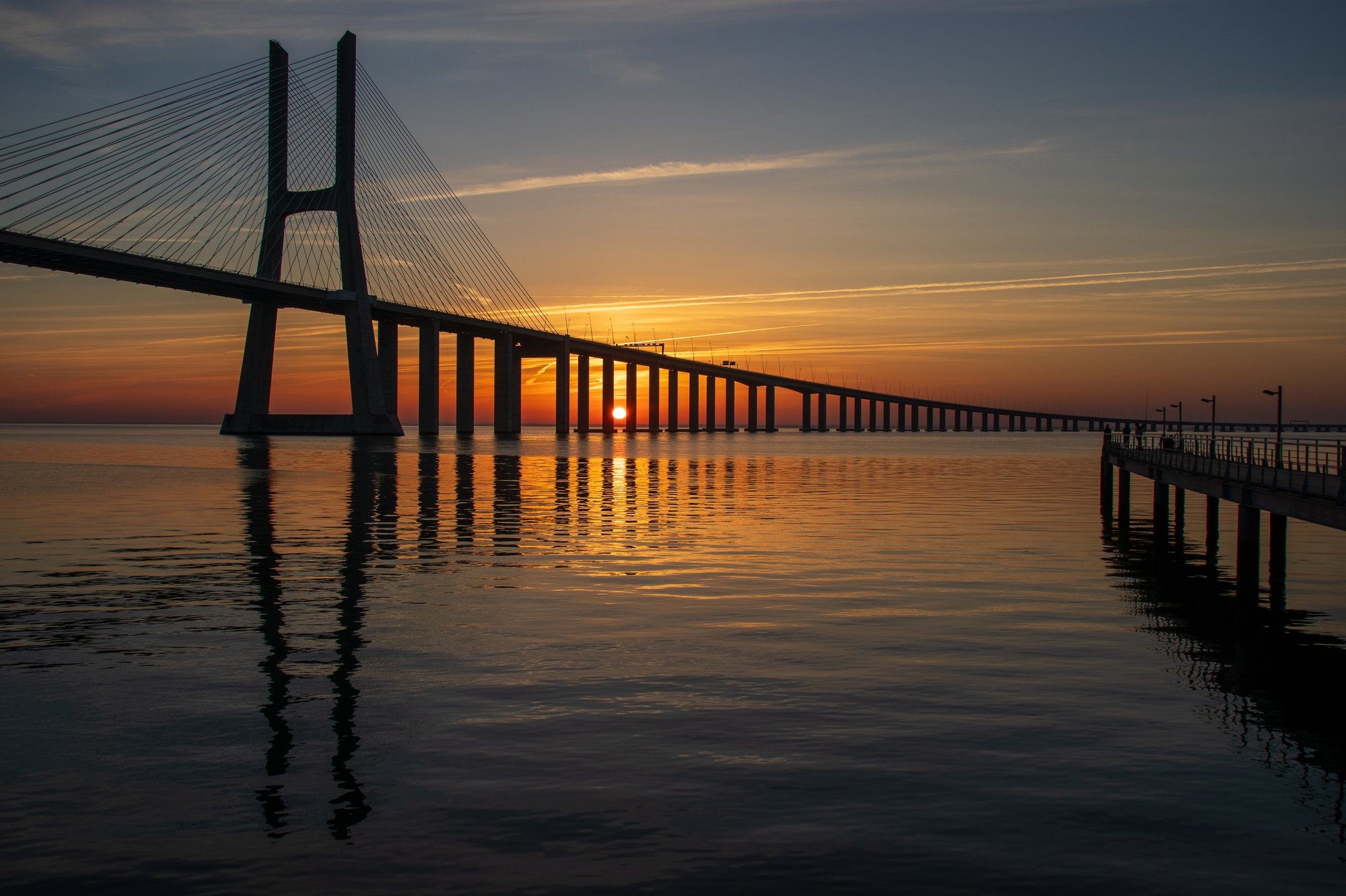 Vasco da Gama Bridge silhouetted against a golden sunrise, with a wooden fishing pontoon reaching out over the Tejo river