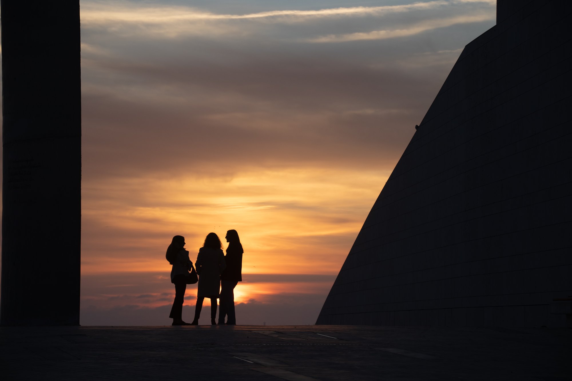 Three women standing in silhouette against a golden sunset, framed between two dark architectural shapes in Belém, Lisbon