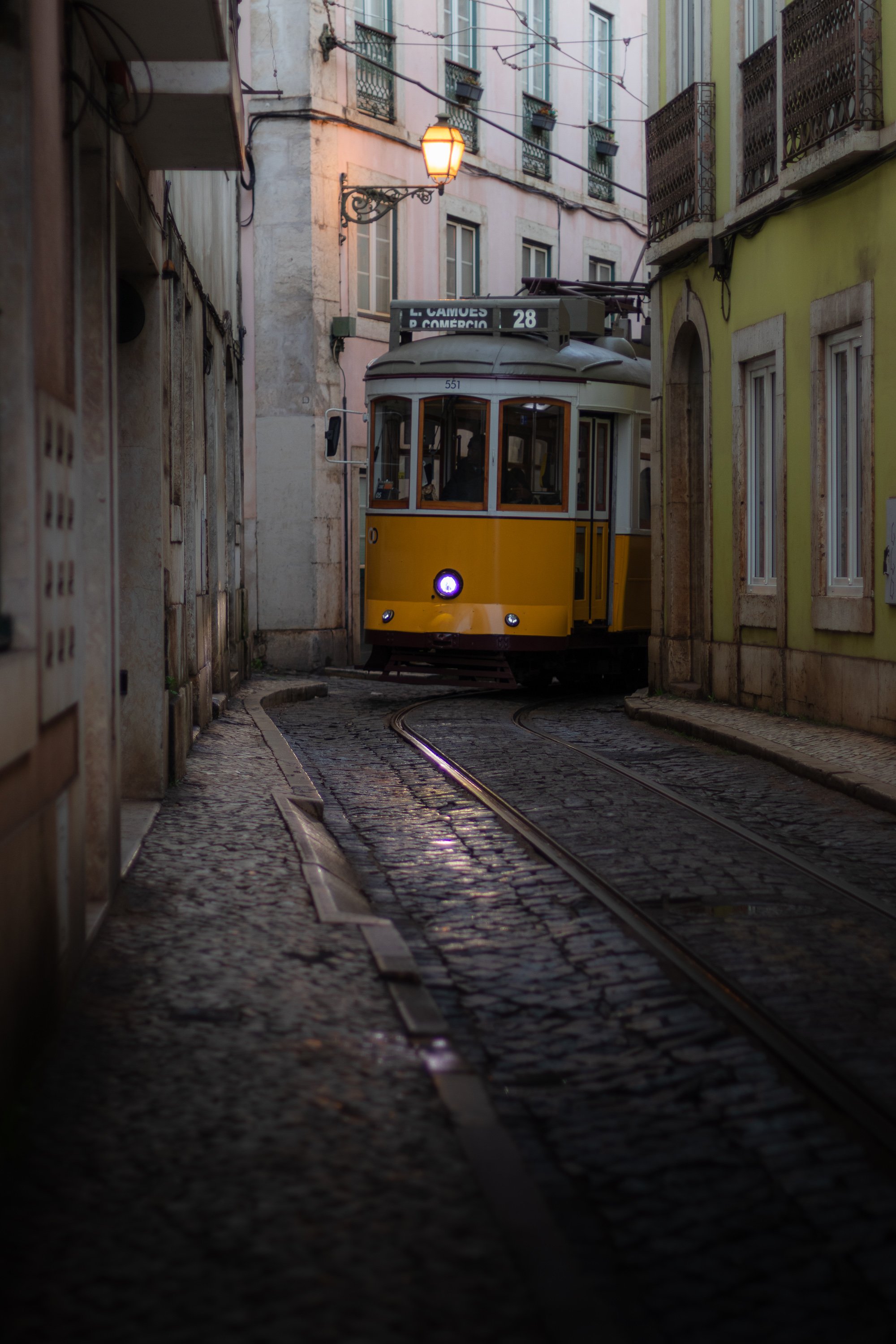 The yellow Tram 28 on a narrow cobbled street in Alfama, Lisbon, with a lit street lamp between pastel buildings