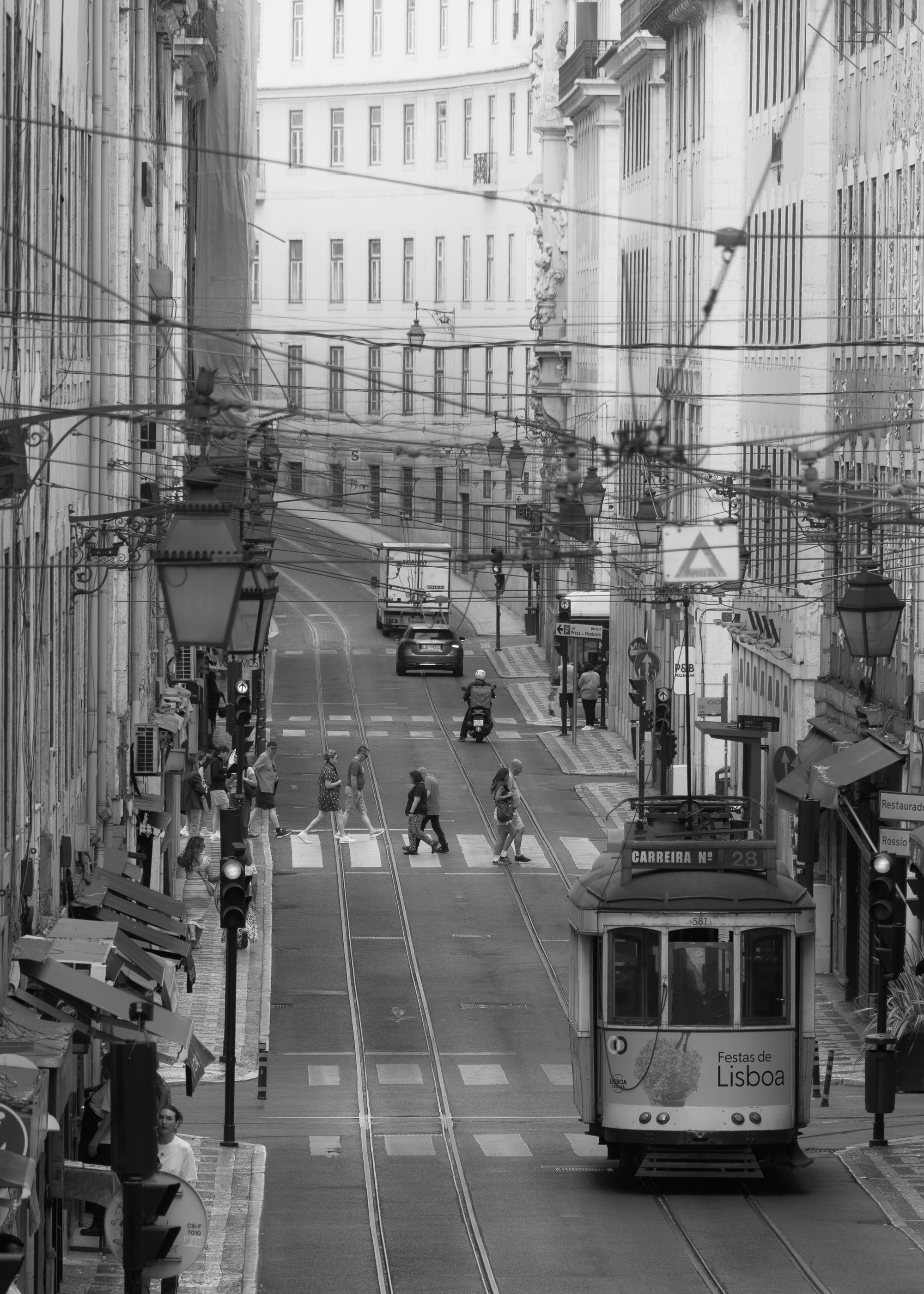 The number 28 tram climbing a cobbled street in Baixa of Lisbon in black and white, with pedestrians crossing ahead.