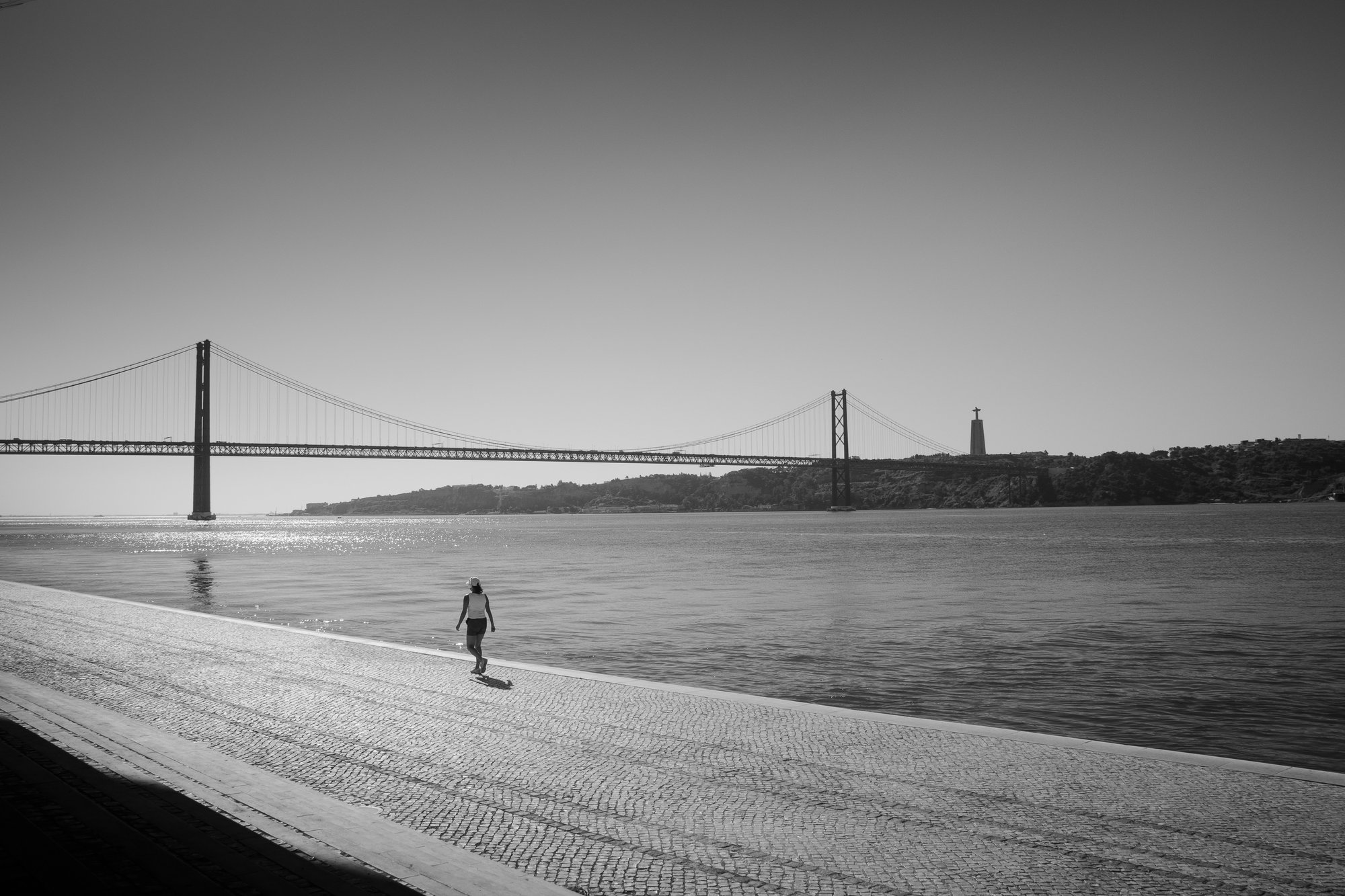 A solitary figure walking along the Tagus riverside in Lisbon, with the 25 de Abril Brisge and Cristo Rei in the distance.