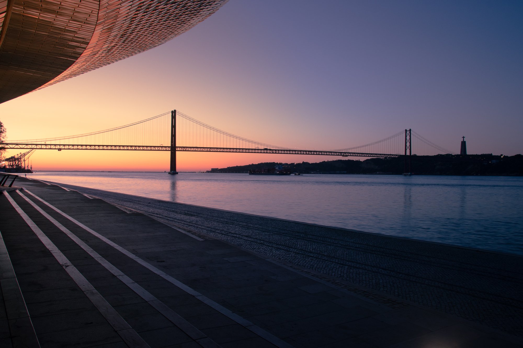 The 25 de Abril Bridge in silhouette at sunrise from Belém, framed by the curved roof of the MAAT and the glowing Tagus River